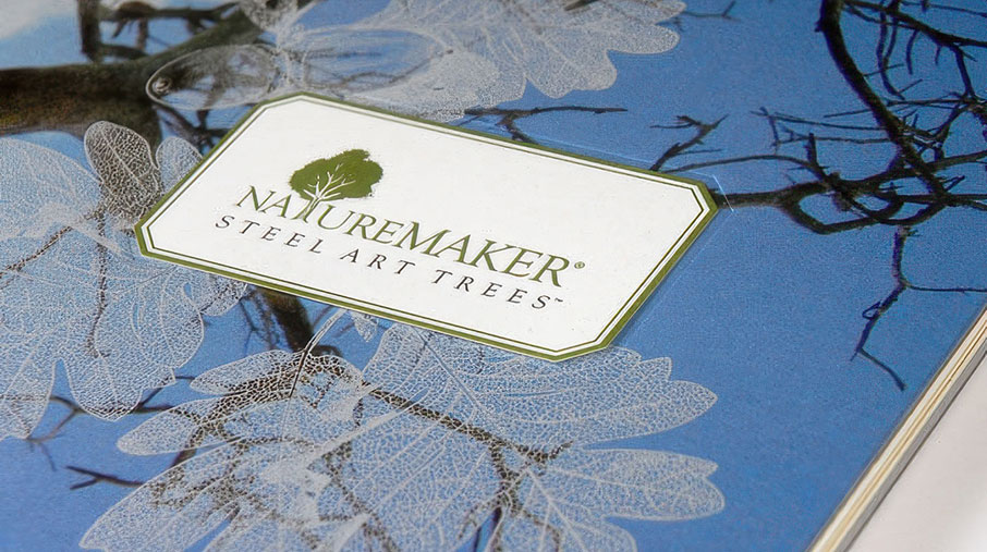 Close-up of the NatureMaker brochure cover featuring the logo “NatureMaker Steel Art Trees” centered in a white label with a green border. The background shows delicate, translucent leaf illustrations and dark tree branches set against a deep blue sky, evoking a blend of nature and art.