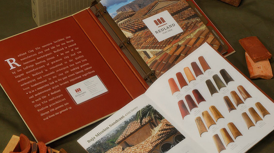 A display of Redland Clay Tile marketing materials featuring a product binder, a printed brochure, and clay tile samples. The open brochure shows various red, orange, and brown roof tile swatches alongside an image of a tiled roof. A wooden crate with curved tile samples and individual tile pieces are arranged on a dark green fabric backdrop, emphasizing natural tones and craftsmanship.