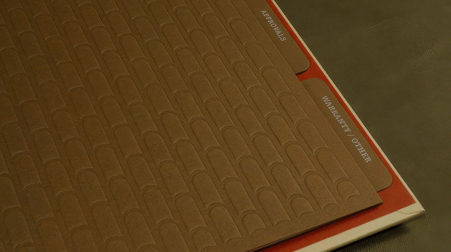 Close-up of a Redland Clay Tile binder with embossed dividers featuring a repeating clay tile pattern. Two labeled tabs are visible—“APPROVALS” and “WARRANTY / OTHER”—protruding from behind the textured brown cover, which rests on a red divider. The background is a muted, dark surface that complements the earthy tones.