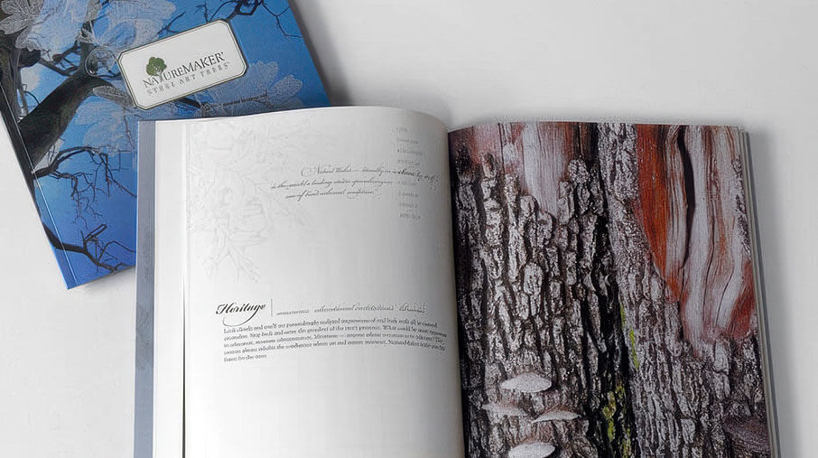 Open NatureMaker brochure showing a detailed close-up of textured tree bark with shades of brown, gray, and red on the right page. The left page features an elegantly typeset description under the heading “Heritage,” with subtle botanical illustrations in the background. A closed brochure with the NatureMaker “Steel Art Trees” logo and a blue floral cover design lies partially visible in the background.