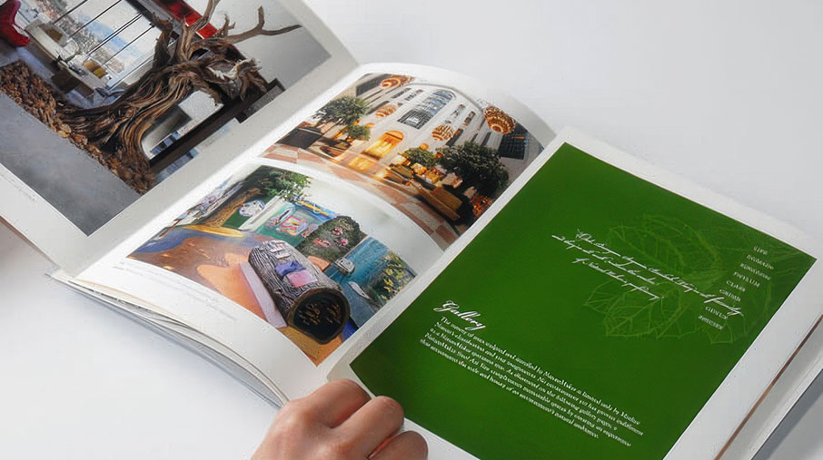 A person flips through a NatureMaker brochure showing a spread of full-color images and a green feature page. The left side displays photos of large-scale artificial tree installations in commercial interiors, including a hotel lobby and themed environments. The right page features a bold green background with leaf outlines, a cursive signature, and a company gallery description in white text.