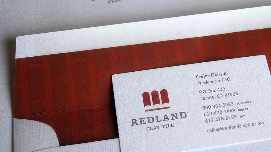Close-up of Redland Clay Tile branded stationery featuring a textured business card and folder. The business card displays the company’s logo—three red clay tiles above the words “Redland Clay Tile”—along with contact information for Carlos Elias, Jr., President & CEO. The folder beneath shows a subtle red tile pattern and complements the card’s clean, embossed design.