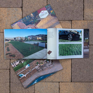 Angelus Paving Stones brochure open to pages highlighting residential applications. The visible spread features a permeable paver driveway with green grass joints and a modern car, alongside a landscaped front yard with paver accents. The closed brochures beneath display an outdoor patio with poolside seating and a front cover labeled "Outdoor Living." All items are placed on a blended tan and brown paving stone surface.