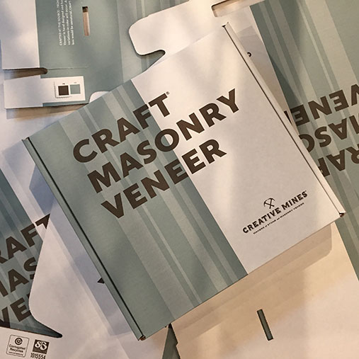 Square packaging box from Creative Mines labeled 'Craft Masonry Veneer' in bold brown text over a minimalist blue-green striped background. The closed box is placed atop several flattened, unfolded versions of the same packaging design, creating a layered effect. The Creative Mines logo appears in the bottom right corner of the box.