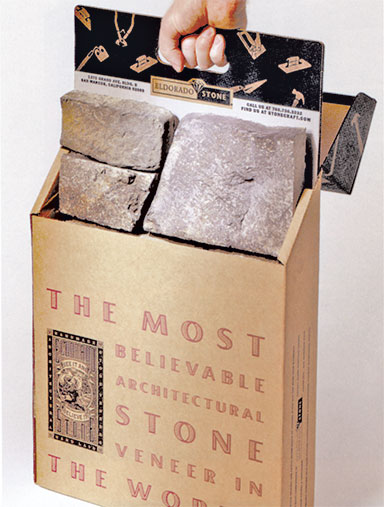 Hand lifting a sample box of Eldorado Stone architectural veneer. The kraft-colored box displays the slogan 'The Most Believable Architectural Stone Veneer in the World' in bold red text, along with the Eldorado Stone logo. Several realistic stone veneer samples are visible inside the box, and a black branded flip panel at the back features product information and illustrations.