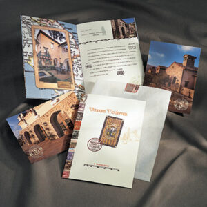A collection of printed marketing materials laid out on a gray fabric backdrop. The materials feature European-inspired architectural imagery with rustic stone homes, ornate doors, and map graphics. The main folder cover reads “Voyages Modernes” in a decorative typeface and includes a vintage-style stamp graphic, evoking a travel or heritage theme.