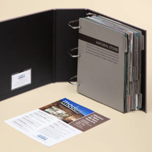 Open binder from Modern Builders Supply featuring a thick set of tabbed reference sheets labeled 'Natural Stone.' The binder cover interior includes a business card holder with a branded Modern Builders Supply card. A promotional flyer with images and text lies in front of the binder on a beige background.