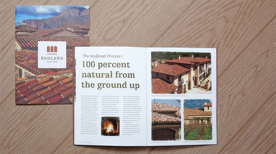 Open Redland Clay Tile brochure on a wood surface, accompanied by a closed cover version showing red clay rooftops and mountains. The open spread features the headline “100 percent natural from the ground up,” with a description of the Redland process on the left page. The right page displays four images of homes with red clay tile roofs, highlighting different architectural styles and details.