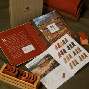 A display of Redland Clay Tile marketing materials featuring a product binder, a printed brochure, and clay tile samples. The open brochure shows various red, orange, and brown roof tile swatches alongside an image of a tiled roof. A wooden crate with curved tile samples and individual tile pieces are arranged on a dark green fabric backdrop, emphasizing natural tones and craftsmanship.
