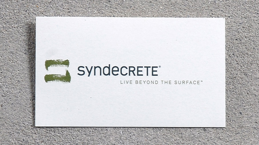 Minimalist Syndecrete business card placed on a textured concrete surface. The card features the Syndecrete logo with two green brushstroke-like marks, followed by the brand name in lowercase black text. Beneath the logo is the tagline 'Live Beyond The Surface™' in light gray.