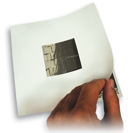 Close-up of a person’s hand lifting the cover of a minimalist square brochure. The cover features a centered photo of concrete tiles in shadow with the words 'LIVE BEYOND' subtly printed. The background is pure white, and the hand’s motion reveals part of the next page underneath.