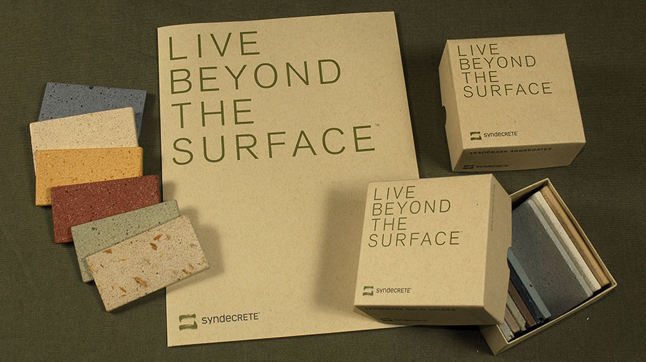Syndecrete presentation kit displayed on an olive green surface. The set includes a large kraft folder labeled 'LIVE BEYOND THE SURFACE' in green text, two branded boxes, and an open box showing neatly stacked rectangular material swatches in various neutral tones. Several loose color and texture samples are arranged to the left, highlighting finishes in blue, cream, gold, red, green, and terrazzo-style beige. The design reflects a natural, minimalist aesthetic.