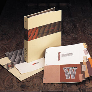 A product binder and set of printed marketing materials for US Tile. The beige binder features a photo of red clay roof tiles and the phrase 'Real Clay Roofs' printed on the spine and cover. Surrounding the binder are sample pages and tabbed inserts, including a prominently visible 'Warranty' page with a large 'W' graphic. The materials are arranged on an ornate, dark wood surface.