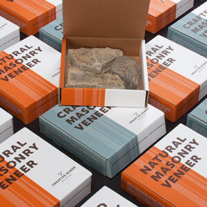 Display of neatly arranged packaging boxes labeled 'Crafted Masonry Veneer' and 'Natural Masonry Veneer' in alternating orange and blue designs. One open box in the center reveals natural stone veneer samples inside. All boxes are branded with the Creative Mines logo and are arranged on a black surface for a clean, modern presentation.