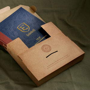 Embossed navy-blue Enkeboll catalog titled 'Architectural Woodcarvings' partially pulled out of a kraft paper presentation box. The box is custom-fitted with a flap closure and features a debossed Enkeboll seal and the text 'Architectural Woodcarvings Catalog' printed in black. The set rests on an olive green fabric background, conveying a sense of craftsmanship and elegance.