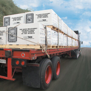 Flatbed semi-truck loaded with white boxed pallets of stone veneer from Eldorado Stone. Each box is labeled 'The Most Believable Architectural Stone Veneer in the World' in bold black text. The truck is driving on a rural road with a grassy hillside on one side and a blue sky with scattered clouds in the background.