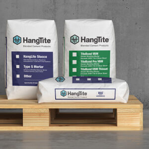 Three bags of HangTite Blended Cement Products displayed on a wooden pallet in front of a concrete wall. The upright bags are labeled with HangLite Stucco Type S Mortar in blue and TiteBond VBM and Thinset products in green. A horizontal bag in front is branded with the HangTite and MAC logos. The packaging uses clean, color-coded design for easy product identification.