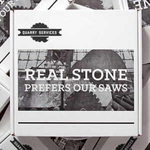 Square white shipping box from Quarry Services with a bold black-and-white label that reads 'REAL STONE PREFERS OUR SAWS.' The label features a high-contrast image of large industrial saw blades cutting through stone. Multiple similar boxes are partially visible beneath, arranged in a layered pattern.