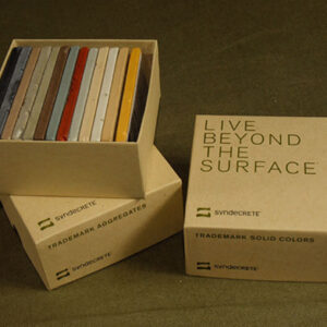 Syndecrete sample packaging displayed on an olive green fabric surface. Three kraft paper boxes are shown—one open, revealing a set of rectangular color swatches in various hues including gray, red, orange, and cream. The boxes are labeled 'Trademark Aggregates,' 'Trademark Solid Colors,' and 'Live Beyond The Surface,' all featuring the Syndecrete logo and minimalist design.
