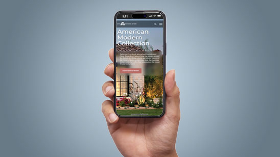Person holding a smartphone displaying the mobile version of the AAA Natural Stone website, featuring the "American Modern Collection" with a large background image of a landscaped outdoor space and a red call-to-action button.