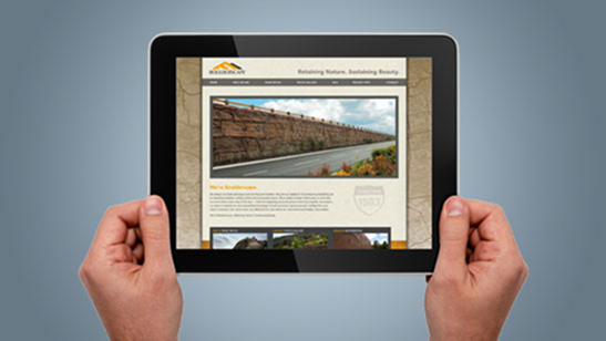 Hands holding a tablet displaying the Boulderscape website, showing a large image of an engineered retaining wall along a roadway with the tagline “Retaining Nature. Sustaining Beauty.”