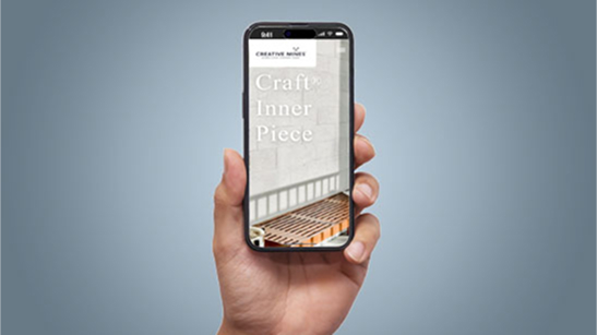 Hand holding a smartphone showing the Creative Mines mobile homepage with the text “Craft Inner Piece” over a minimalist architectural background featuring a bench and a concrete wall.