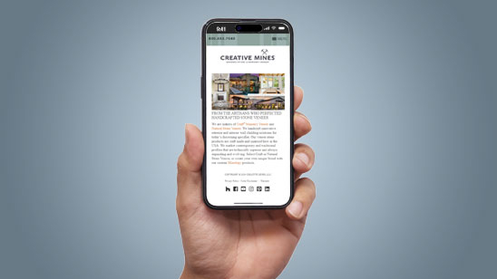 Hand holding a smartphone displaying the Creative Mines mobile website, featuring an article about a product appearance on Property Brothers, with images of modern homes and stone veneers above the text.