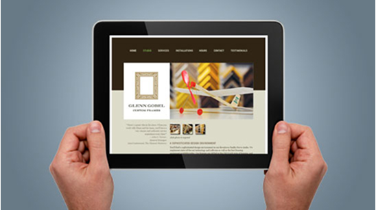 Hands holding a tablet displaying the Glenn Gobel Custom Frames website, showcasing an image of picture frame corners and tools, with navigation links for home, studio, services, installation, and more.