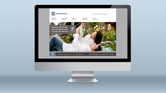 Desktop monitor displaying the Headwaters website homepage, featuring a banner image of a smiling woman and child lying in a landscaped yard with the tagline “Improving Lives through Innovative Advancements in Construction Materials.”