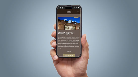 Hand holding a smartphone displaying the Modern Builders Supply mobile website, featuring a welcome message and an image of the storefront with a blue “modern” sign illuminated at night.