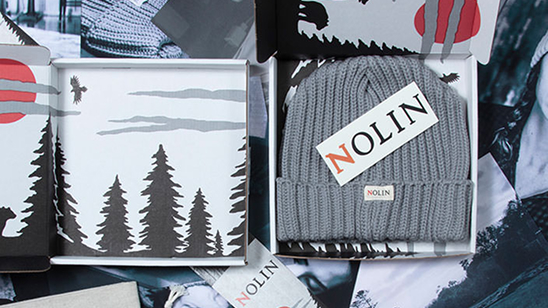 Nolin brand packaging featuring a gray knit beanie with a Nolin tag, presented in a box with forest-themed artwork. The logo displays the word “NOLIN” in serif type, with the letter “N” highlighted in red.