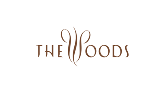 The Woods logo in elegant brown lettering, featuring a stylized, nature-inspired “W” with sweeping curves that resemble tree branches or vines.