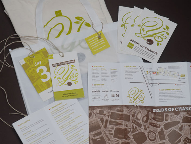 A flat lay display of branded materials from the AIGA Y13 Conference themed “Seeds of Change.” Items include printed schedules, speaker lists, maps, name badges, brochures, and tote bags, all featuring the Y13 logo with leafy graphics and a seed illustration. The consistent color palette of green, brown, and white reinforces the nature-inspired conference theme. Tags with twine and eco-conscious design elements emphasize sustainability and creativity.