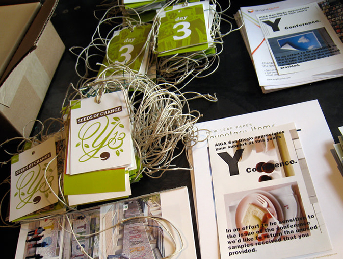 A preparation table for the AIGA Y13 Conference, showcasing stacks of attendee badges, string lanyards, and informational handouts. The badges display the Y13 “Seeds of Change” logo in green and brown, along with “day 3” labels. Printed materials emphasize sustainability and recycling, with one sheet explaining AIGA’s effort to reuse samples returned by attendees. The overall scene reflects an eco-conscious and well-organized event setup.