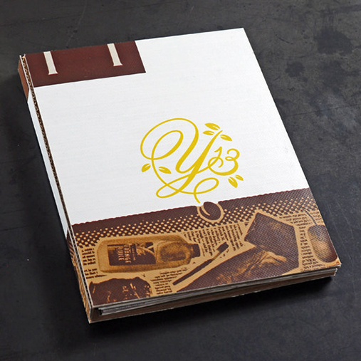 A closed conference binder or portfolio from the AIGA Y13 Conference. The cover features the stylized yellow “Y13” logo with leaf accents on a clean white background. The bottom portion includes a textured brown design with illustrations of sustainable materials like bottles, paper, and utensils, emphasizing the event's “Seeds of Change” theme and focus on eco-conscious design. The binder rests on a dark surface.