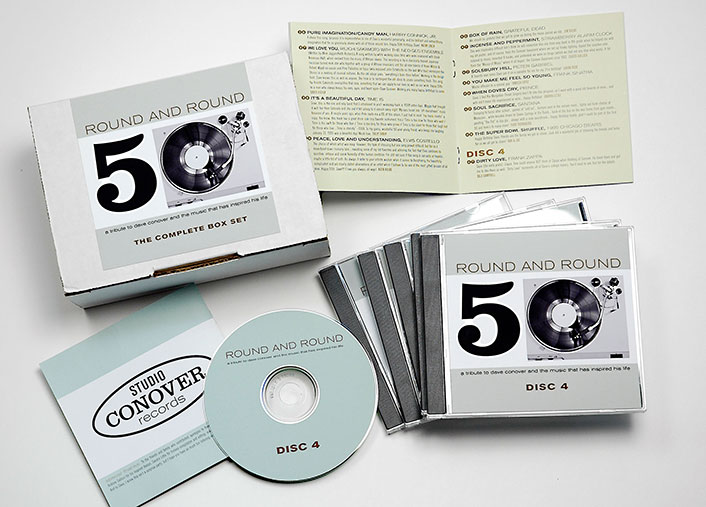 Box set titled “Round and Round 50 – The Complete Box Set” by Studio Conover Records, displayed with five CD cases, a lyric booklet, and Disc 4 shown separately. The packaging design features a retro record graphic with minimalist typography in black, white, and pale mint green. The booklet lists classic song titles from various decades, suggesting a nostalgic music collection.