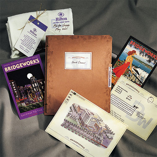Promotional materials for BridgeWorks displayed on a soft fabric surface. The collection includes brochures, vintage-style postcards, a site plan illustration, a copper-toned folder labeled with “David Conover,” and a Hilton-branded towel bundle from a Las Vegas event. The design aesthetic blends retro travel imagery with detailed hospitality marketing content.