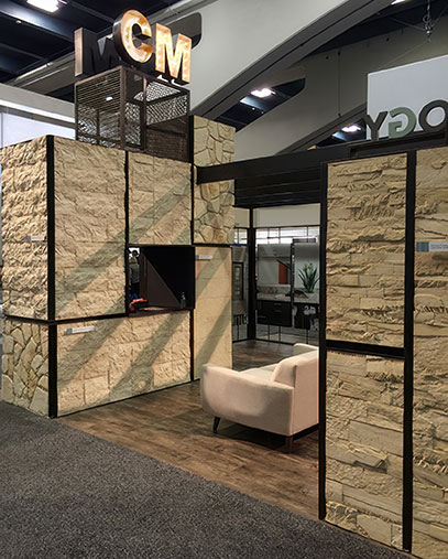 A Creative Mines trade show booth designed with large stone veneer panels in varying textures and tones. The structure includes black-framed divisions and a wood-style floor, with a cream-colored modern lounge chair inside. A small monitor is embedded in one of the walls. At the top, illuminated initials “CM” are mounted above a metal mesh structure, serving as a branded identifier visible from afar. The surrounding booths and signage hint at a bustling convention environment.