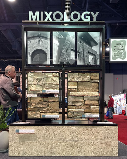 A trade show display booth by Creative Mines titled “MIXOLOGY,” featuring a tall structure with black metal framing. The top section showcases three black-and-white architectural photographs of stonework. Below are four side-by-side panels displaying different textured stone veneer styles in natural earth tones. The base of the structure is clad in a cream-colored textured stone panel. Visitors are seen interacting nearby, and the surrounding environment includes other vendor booths and red carpeted flooring. The display emphasizes product variety and stone craftsmanship.