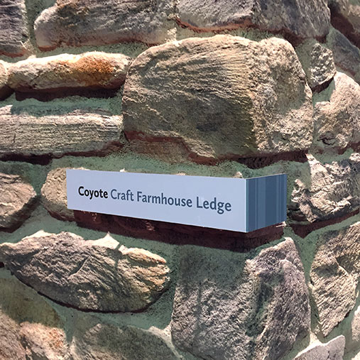 A close-up of a stone veneer display at a trade show, labeled “Coyote Craft Farmhouse Ledge.” The sign is mounted directly onto the stone wall and features a clean, modern design with the name in black and gray text, along with a vertical stripe graphic on the right side. The stone itself has a rugged, irregular texture with varied shades of brown, gray, and beige, showcasing the product's natural, handcrafted appearance.