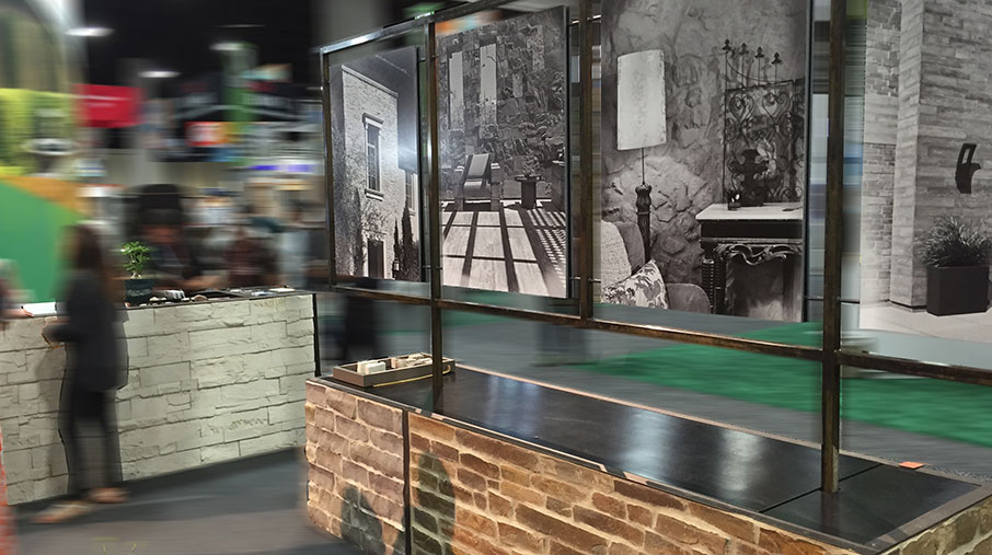 A trade show booth featuring Creative Mines stone veneer products. The display includes textured stone surfaces on the counters and lower walls, with a steel-framed partition holding three large black-and-white lifestyle photographs showcasing stone in architectural and interior design settings. The background is slightly blurred, capturing the energy of the exhibition space with attendees walking by and other booths visible. The booth’s design emphasizes craftsmanship, texture, and sophisticated stone applications.