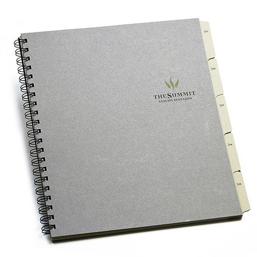 Spiral-bound presentation book for The Summit at Rancho Bernardo, created by Cruzan. The cover is a minimalist gray with the logo and title printed in green and black. Visible tab dividers labeled “01” through “06” extend from the right edge, indicating organized sections within the document.