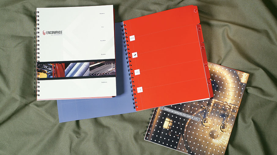 A set of spiral-bound proposal documents for PacRim Mechanical's Encompass division. One binder has a clean white cover with the Encompass logo and labeled sections such as “Fire,” “Facilities,” and “Solutions.” Inside are bold red tab dividers with white icons and section labels. A second booklet with a glossy cover features an industrial photograph of metallic pipes and valves overlaid with a grid of white dots. All materials are displayed on a green fabric surface, showcasing a professional and cohesive branding system.