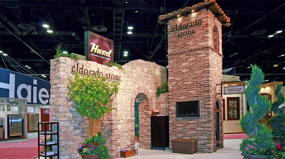 A large, immersive trade show booth for Eldorado Stone designed to resemble a rustic Mediterranean villa. The structure includes stone and brick facades, arched doorways, and a tall tower with a clay tile roof. The Eldorado Stone logo is prominently displayed on the stone wall and tower. Lush greenery and potted plants decorate the booth, creating an inviting and natural atmosphere. A flat-screen TV and product signage are integrated into the design, while nearby booths and overhead signage from other brands are visible in the background.