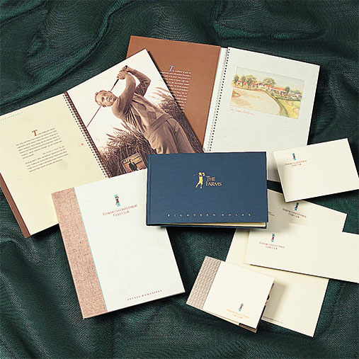 Branded print materials for The Farms, displayed on a dark green mesh fabric. The collection includes brochures, stationery, and a navy blue directory book with gold lettering and logo. Open booklets reveal a black-and-white photo of a golfer in mid-swing and an illustrated golf course scene, emphasizing a refined, upscale golf community identity.