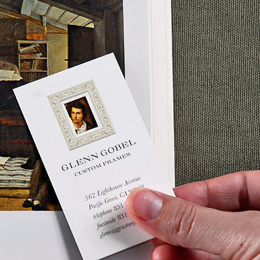 A hand holds a business card for Glenn Gobel Custom Frames in front of a framed painting. The card features a miniature classical portrait inside a decorative frame, along with the business name, contact details, and address in Pacific Grove, California. The background includes a partial view of a textured green wall and a corner of the framed artwork.