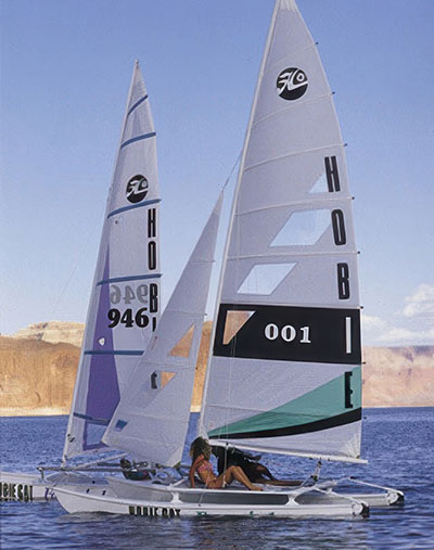 Two Hobie catamarans sailing side by side on a calm lake, with clear blue skies and desert cliffs in the background. The sails prominently display the Hobie logo and sail numbers, while two people relax on one of the boats, enjoying the water and sun.