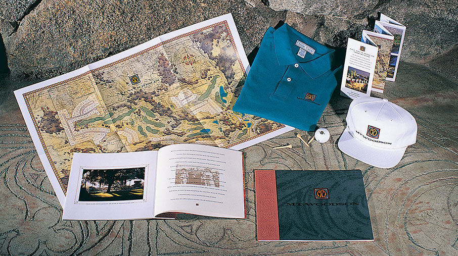 A display of branded promotional materials for Mt. Woodson Golf Club, arranged on a stone surface. Items include a detailed illustrated course map, an open photo book, a dark green and red brochure with the Mt. Woodson logo, a teal polo shirt and a white baseball cap embroidered with the logo, golf tees, a logo golf ball, and several folded brochures. The materials convey a premium, nature-inspired golfing experience with a focus on branding consistency and elegance.