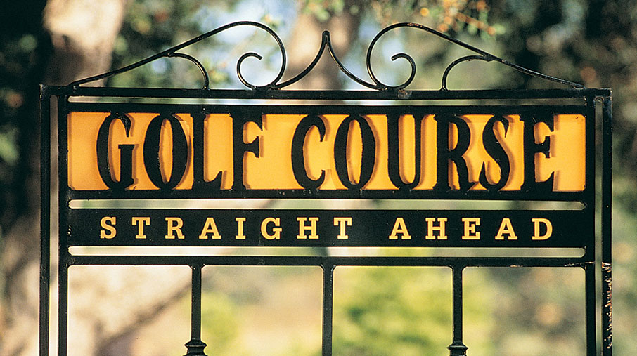 A wrought iron directional sign at Mt. Woodson Golf Club with decorative scrollwork at the top. The sign features bold, vintage-style yellow and black lettering that reads “GOLF COURSE” on the top panel and “STRAIGHT AHEAD” on the panel below. The background is softly blurred with trees and sunlight, emphasizing the rustic and inviting atmosphere of the course.