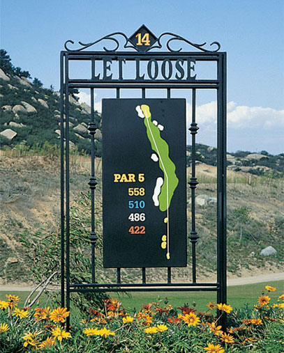 A decorative golf course hole marker for Mt. Woodson Golf Club, displaying hole number 14 with the name “LET LOOSE” in elegant wrought-iron lettering. The central sign shows a colorful map of the par 5 hole layout with yardages listed: 558, 510, 486, and 422. The sign is surrounded by wildflowers in the foreground, with rolling hills, rocks, and a path in the background under a partly cloudy sky.